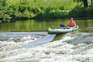 Ein Bootsfahrer mit oranger Rettungweste und Paddel steht mit seinem grünen Kanu am Anfang der Bootstreppe in der Altmühl. Das Wasser rauscht hier an dem steinernen Wehr hinunter. Die Bootsrutsche ist eine Vertiefung für die Fische und auch Boote. Das Ufer ist sattgrün auf dem die Sonne scheint.