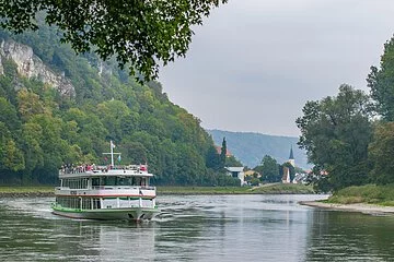 Ein Personenschiff fährt auf dem Main-Donau-Kanal entlang. Links ist das Ufer steil und mit Bäumen bewachsen, rechts geht es flacher mit einem kleinen Kiesrand ins Wasser. Am Horizont erkennt man einen Kirchturm in der Ferne.