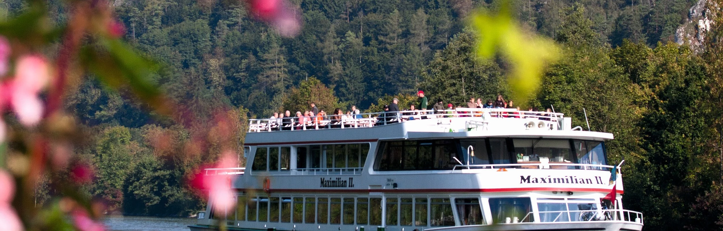 Ein voll besetztes Personenschiff auf dem Main-Donaukanal. Hinter ihm ragen die Hänge auf. Links blitzt ein grüner Zweig mit rosa Blüten ins Bild.