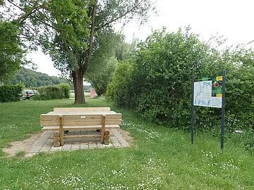 Bootsrastplatz Inching Die Sitzgelegenheit aus Holz auf gepflastertem Grund ist inmitten einer grünen Wiese am Uferrand mit Büschen und Bäumen. Daneben steht eine Infotafel zum Thema Bootwandern.