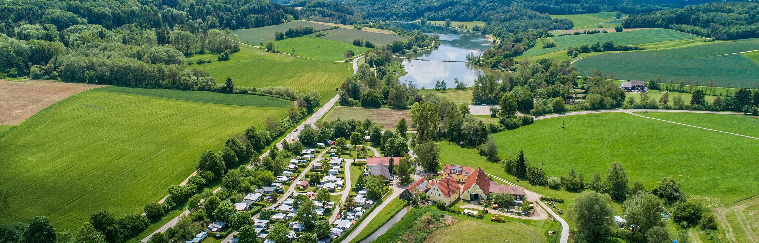 Der Campingplatz Hasenmühle von oben inmitten grüner Wiesen und Bäumen. Hinter dem Platz ist der Hahnenkammsee zu sehen.