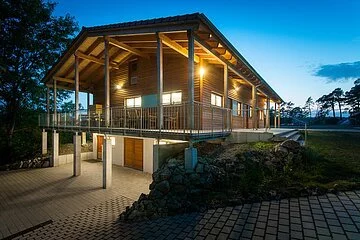 Ein Holzhaus mit großer Terrasse ringsherum ist hellbeleuchtet in der Nacht. Ein gepflasteter Weg führt links hinunter zum selben Gebäude, das im Untergeschoss gemauert und im Hang gebaut ist. Die Terrasse wird durch Pfeiler gestützt.