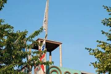 Eingerahmt von zwei großen Bäumen steht ein Aussichtsturm mit einem Kunstobjekt auf grünem Hintergrund. Links ist eine Fahne gehisst.