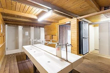 Der Waschraum hat zwei große Waschbecken in der Mitte . Rechts geht es zu den Duschen und WC's. Das Gebäude ist aus Holz gebaut.