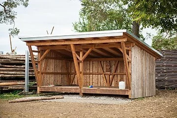 Eine Schutzhütte aus Holz steht vor einem Holzzaun. Dahinter stehen einige Bäume.