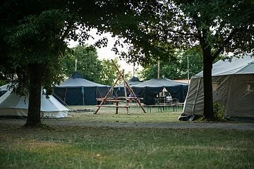 Auf dem Zeltplatz stehen unter Bäumen größere Mannschaftszelte in weiß und blau. In der Mitte ist eine Feuerstelle mit Grillmöglichkeit.