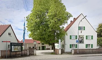 Bauerngerätemuseum Hundszell Der Eingangsbereich des Museums mit seinen Gebäudlichkeiten. Links ist zudem eine Infotafel und eine Fahne angebracht. Der Blick in den Hof zeigt ein weiteres Gebäude und einen großen Baum.