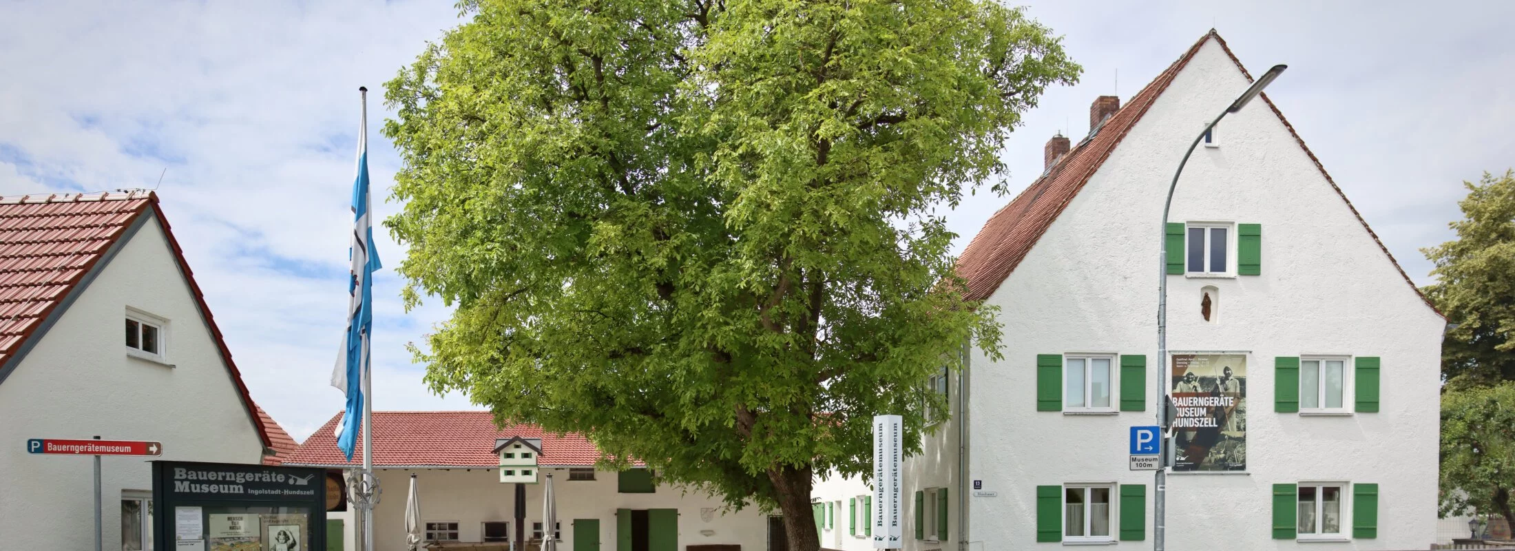 Der Eingangsbereich des Museums mit seinen Gebäudlichkeiten. Links ist zudem eine Infotafel und eine Fahne angebracht. Der Blick in den Hof zeigt ein weiteres Gebäude und einen großen Baum.