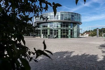 AUDI Museum mobile Das Museum mobile mit seiner ungewöhnlichen, runden Architektur. Vor dem Museum erstreckt sich die gepflasterte Piazza und links ragt ein Baum herein.
