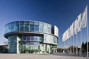 Das Museum mobile mit seiner einzigartigen runden Architektur. Vor dem Museum die Piazza und rechts sind sechs Fahnen gehisst. Der Himmel strahlt wolkenlos.