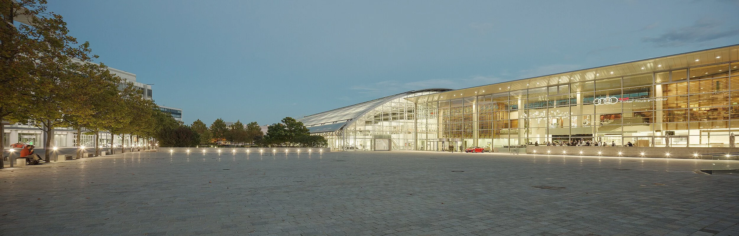 Die Piazza erstreckt sich vor uns und rechts das Audi Forum hell erleuchtet am Abend.