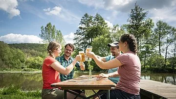 Biergarten des Gasthof Hollerstein Zwei Pärchen sitzen an einer Biertischgarnitur und prosten sich mit gefüllten Biergläsern zu. Im Hintergrund ist die Altmühl und grüne Bäume zu sehen.