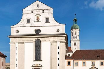 Die Aufnahme zeigt die Fassade der Schutzengelkirche mit einem hohen Fenster über der Eingangstür, rechts daneben liegt das angrenzende Priesterseminar. Direkt vor der Kirche befindet sich der Leonrodplatz. Schräg rechts hinter der Kirche ist ein hoher Turm mit einer Uhr zu erkennen.