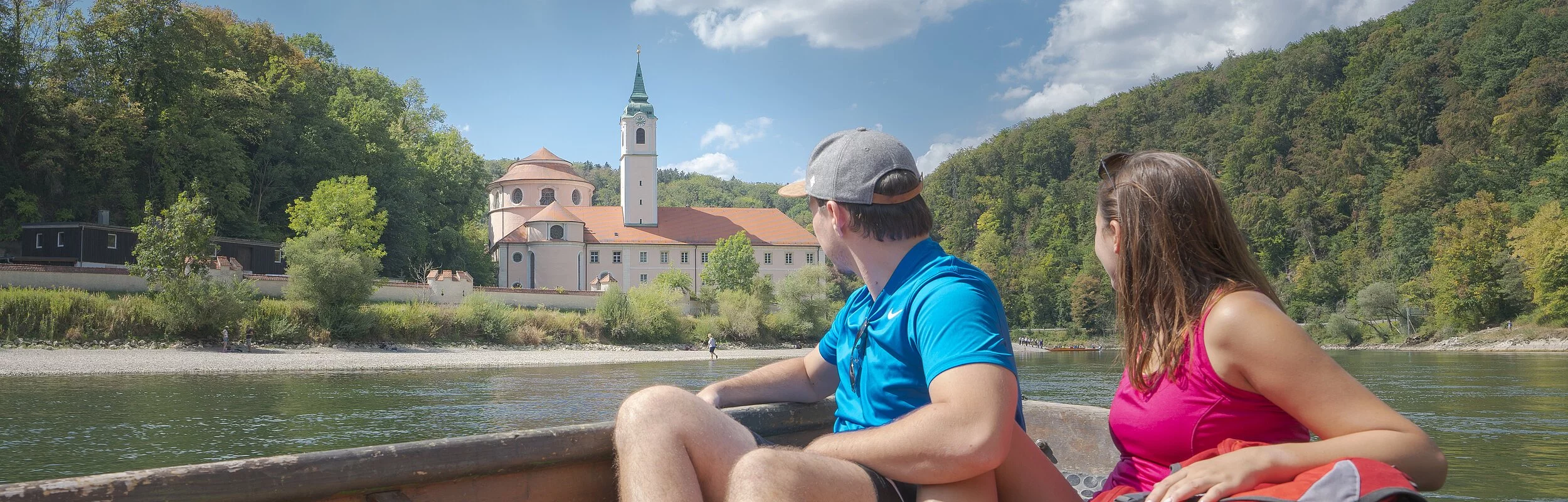 In dem hölzernen Boot sitzen ein Mann und eine Frau, beide in Wanderkleidung mit dem Blick Richtung Kloster Weltenburg gerichtet. Das Kloster ist im Hintergrund von weitem zu sehen umrahmt von grünen Wäldern.