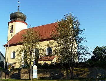 Ein kleine Kirche mit kurzem Kirchenschiff und kleinem Kirchturm mit Zwiebeldach. Die Kirche hat eine gelbe Fassade und ein rotes Ziegeldach. Vor der Kirche stehen zwei große Bäume und eine Steinmauer.