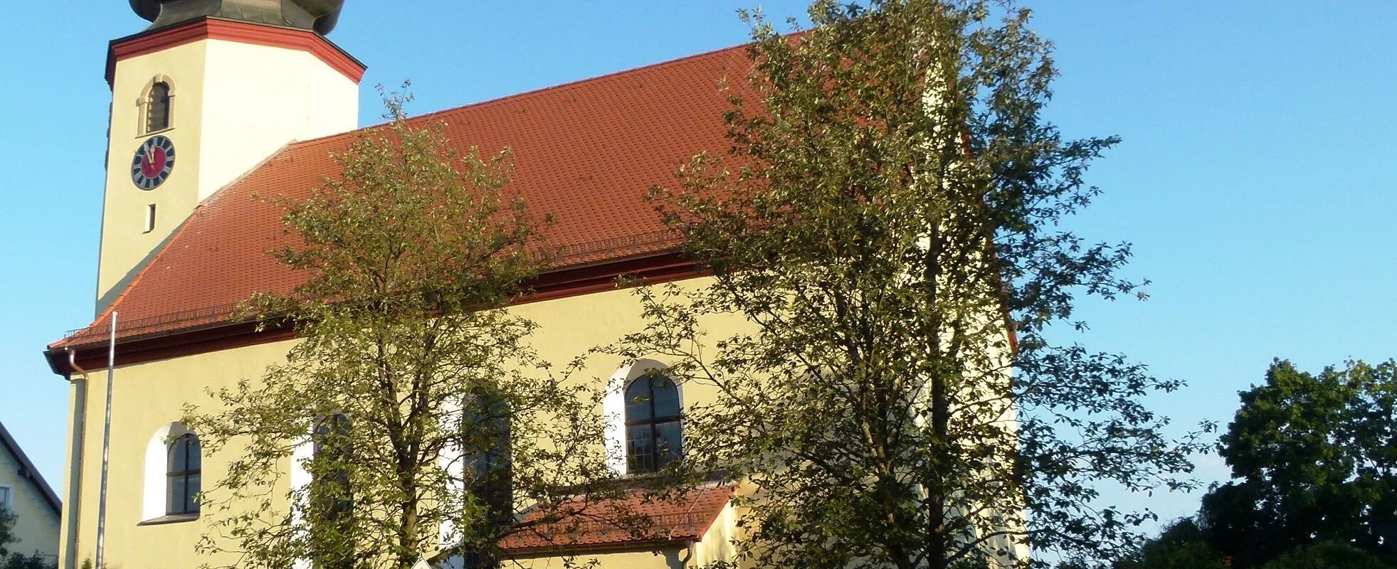 Ein kleine Kirche mit kurzem Kirchenschiff und kleinem Kirchturm mit Zwiebeldach. Die Kirche hat eine gelbe Fassade und ein rotes Ziegeldach. Vor der Kirche stehen zwei große Bäume und eine Steinmauer.
