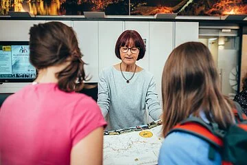 Eine Mitarbeiterin der Tourist-Information berät zwei Kundinnen. Sie hat eine Karte aufgeklappt und erklärt dazu etwas.