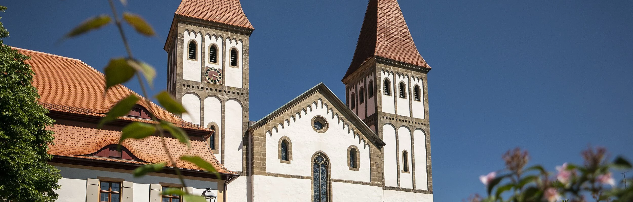 Das Kloster mit seinen zwei Türmen inmitten grüner Büsche. Der Himmel strahlt wolkenlos.
