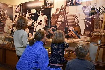 Eine Frau sieht sich mit drei Kindern eine Vitrine im Käthe-Kruse-Puppenmuseum an in der erklärt wird, wie die Puppen hergestellt werden.