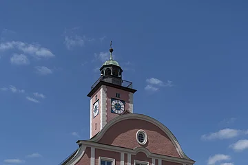 Das Bild zeigt ein prächtiges vierstöckiges Haus mit einem geschwundenen Giebel. Darüber thront im Hintergrund ein Glockenturm mit Ziffernblätter und einem Kupferdach sowie einem Balkon, der um den gesamten Turm führt. Das Rathaus und der Turm sind rotbraun gestrichen. Der Eingang in Form eines zweiflügeligen Holztors befindet sich in der Mitte der Vorderseite, direkt darüber ist ein Balkon zu erkennen, der durch zwei Säulen links und rechts des Eingangstores gestützt wird.