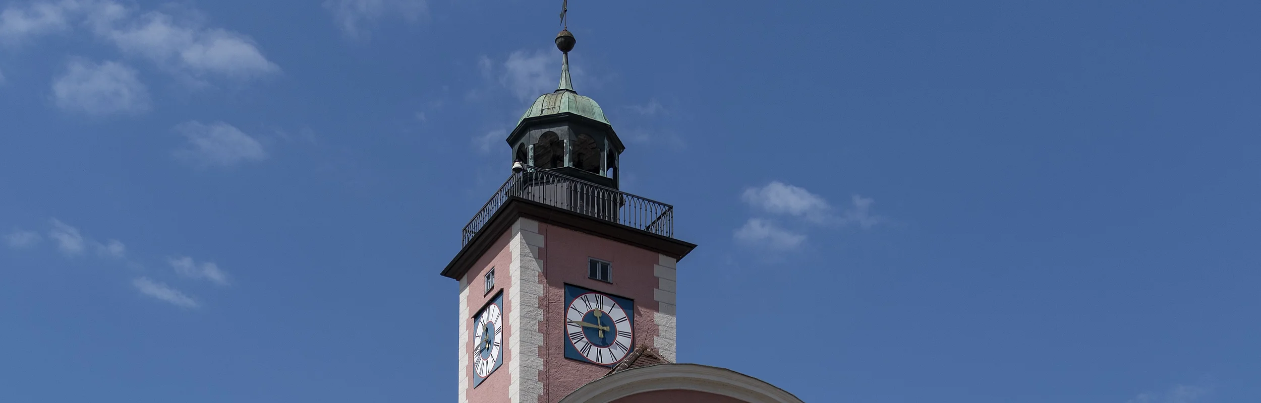 Das Bild zeigt ein prächtiges vierstöckiges Haus mit einem geschwundenen Giebel. Darüber thront im Hintergrund ein Glockenturm mit Ziffernblätter und einem Kupferdach sowie einem Balkon, der um den gesamten Turm führt. Das Rathaus und der Turm sind rotbraun gestrichen. Der Eingang in Form eines zweiflügeligen Holztors befindet sich in der Mitte der Vorderseite, direkt darüber ist ein Balkon zu erkennen, der durch zwei Säulen links und rechts des Eingangstores gestützt wird.