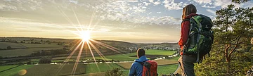 Wanderer auf dem Altmühltal-Panoramaweg genießen den Sonnenuntergang auf dem Burgsteinfelsen Eine Frau mit roter Jacke und grünen Wanderrucksack steht auf dem Burgsteinfelsen, vor ihr sitzt ein Mann in blauer Jacke und roten Rucksack. Beide schauen ins Tal hinunter und genießen den Sonnenuntergang. Über ihnen ziehen die Wolken am blauen Himmel entlang.