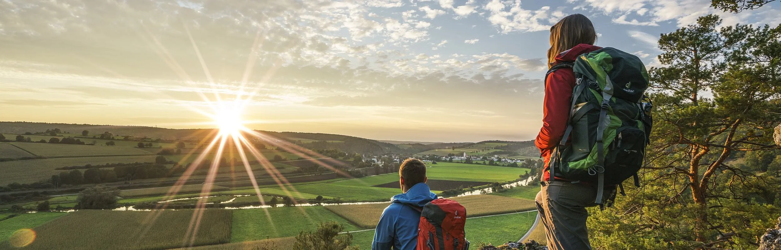 Eine Frau mit roter Jacke und grünen Wanderrucksack steht auf dem Burgsteinfelsen, vor ihr sitzt ein Mann in blauer Jacke und roten Rucksack. Beide schauen ins Tal hinunter und genießen den Sonnenuntergang. Über ihnen ziehen die Wolken am blauen Himmel entlang.
