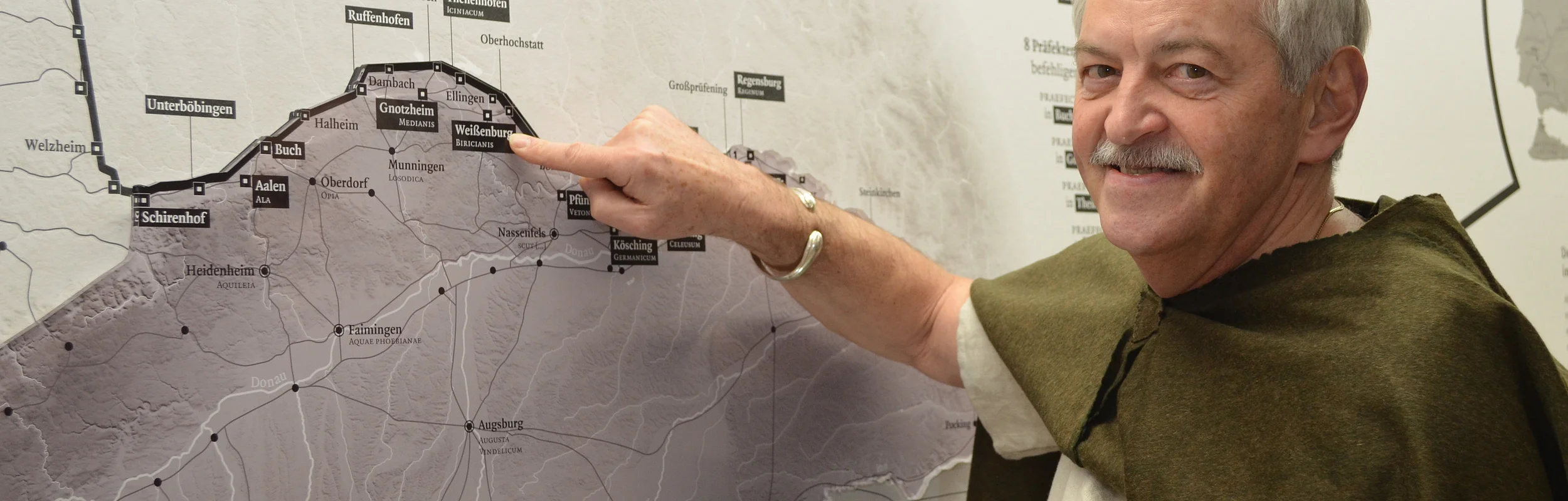 Ein Mann mit einem grünen altertümlichen Umgang schaut lächelnd in die Kamera und zeigt dabei mit seinem Zeigefinger auf eine Landkarte.