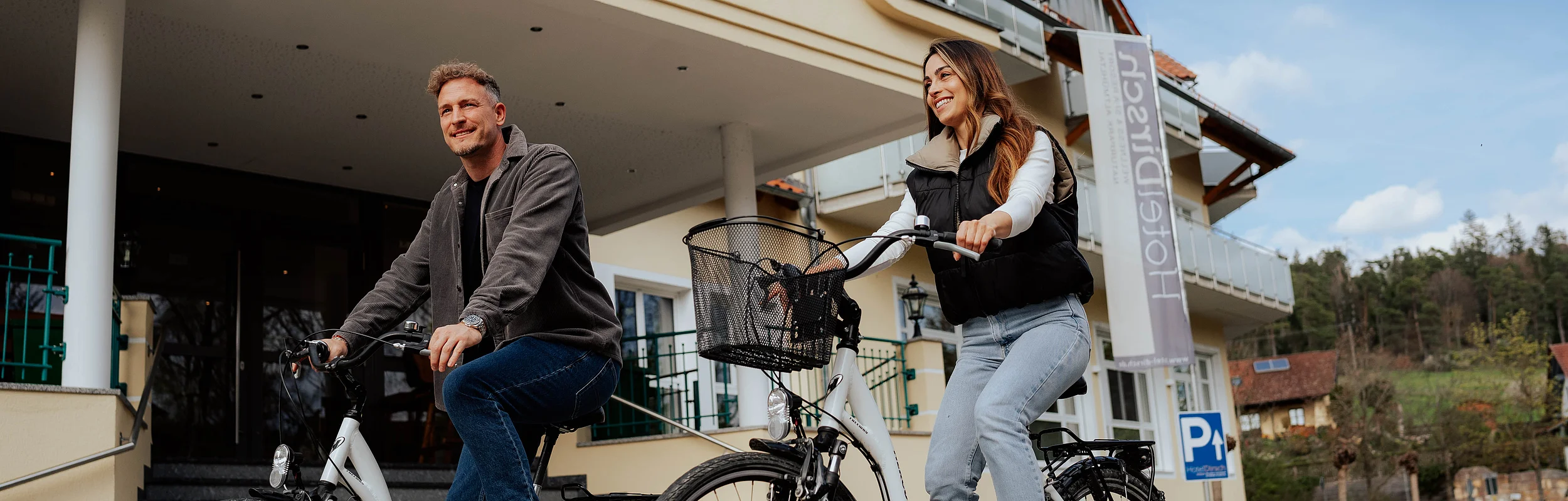 Eine Frau und ein Mann auf dem Fahrrad vor dem Hoteleingang Hotel Dirsch in Emsing.