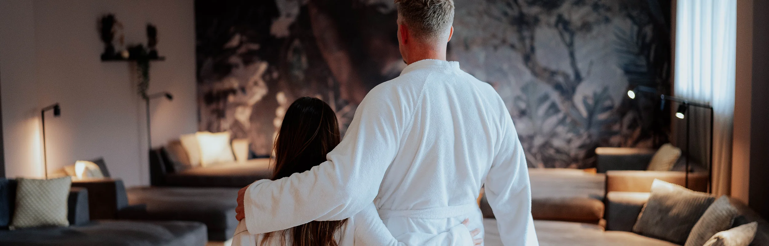 Eine Frau und ein Mann im Bademantel mit Handtücher im Ruheraum des Hotel Dirsch in Emsing. Im Hintergrund Liegen und ein Sofa mit einer kleinen Beleuchtung im Raum.