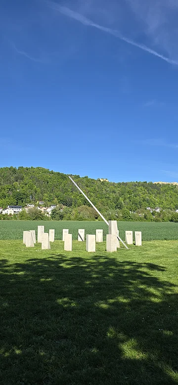 Die Sonnenuhr auf der grünen Wiese ist aus massiven Steinen gebaut. Im Hintergrund ist der Wald und der blaue Himmel zu sehen.