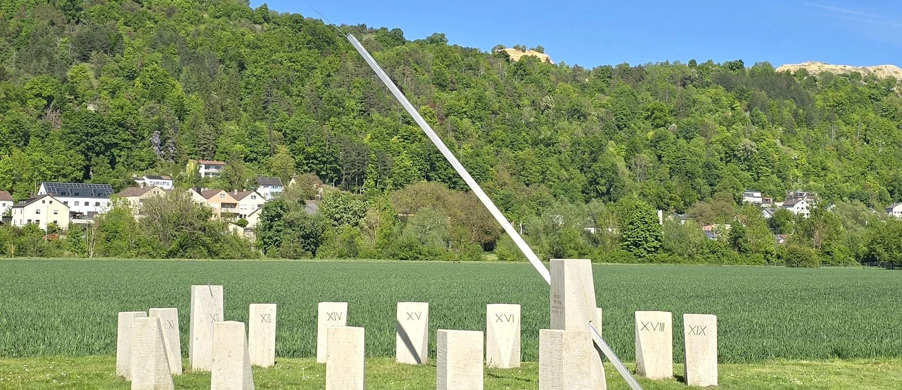 Die Sonnenuhr auf der grünen Wiese ist aus massiven Steinen gebaut. Im Hintergrund ist der Wald und der blaue Himmel zu sehen.