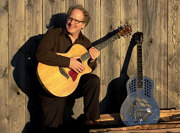 Paul Stowe Ein dunkel gekleideter Mann mit Brille und Gitarre in der Hand. Eine weitere Gitarre lehnt an einer Holzwand.