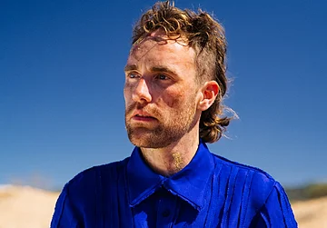 Kaurna Cronin Band Ein Mann mit längerem Haar und blauem Hemd, im Hintergrund blauer Himmel.