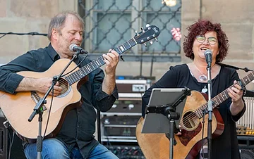 Ein dunkel gekleideter Herr mit Gitarre in der Hand und eine dunkel gekleidete Frau mit Brille, ebenfalls mit Gitarre. Vor beiden steht ein Notenständer und zwei Mikrofone. Im Hintergrund sieht man einen Verstärker stehen und ein vergittertes Fenster.
