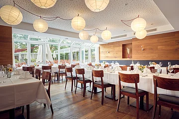 Ein großer Saal mit bodentiefen Glasfenstern, Holzstühlen und Lampions, die von der Decke hängen.