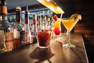 Drei Cocktails sind auf dem schwarzen Tresen platziert, im Hintergrund befindet sich eine Bar.
