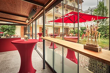 Rote Stehtische sind im Außenbereich platziert. Im Hintergrund sind weitere Sitzmöglichkeiten und ein Sonnenschirm.