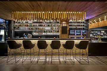 Die hohen Stühle sind mit Blick zur Bar gerichtet aufgestellt. An der Decke sind goldene Röhren angebracht.