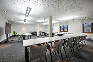 Im kahlen Raum stehen weiße Tische in U-Form-Bestuhlung aufgestellt mit grauen Stühlen.