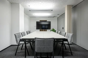 Die grauen Stühle sind um einen weißen langen Tisch mit Blick zum Bildschirm gerichtet. Der Raum ist minimalistisch eingerichtet.