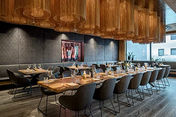 Der große und offene Raum ist mit rustikalen Holztischen und grauen Stühlen versehen. Von der Decke hängen zylinderförmige braune Lampen.