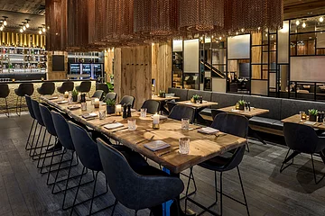 Der große und offene Raum ist mit rustikalen Holztischen und grauen Stühlen versehen, er hat eine ruhige Atmosphäre. Von der Decke hängen zylinderförmige goldene Kettenlampen.