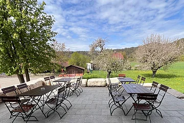 Außenterrasse mit leeren Tischen und Stühlen, umgeben von blühenden Bäumen und grüner Landschaft.