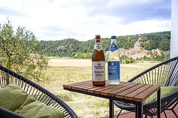 Zwei Flaschen, eine Bier- und eine Wasserflasche, stehen auf einem Holztisch vor einer Landschaft mit Feldern und Bäumen.