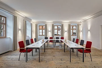 Die weißen Tische sind in U-Form-Bestuhlung platziert. Um sie herum stehen graue und rote Stühle.