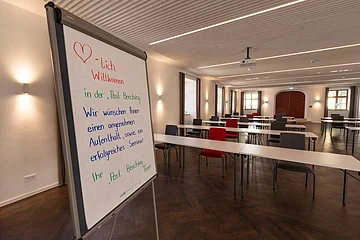 Die Tafel steht vorne, im Raum sind verschiedenfarbige Stühle an weißen Tischen verteilt.