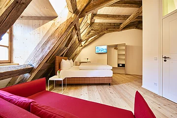 Das Doppelbett befindet sich unter der Dachschräge im Zimmer, die rote Couch steht vorm Fenster.