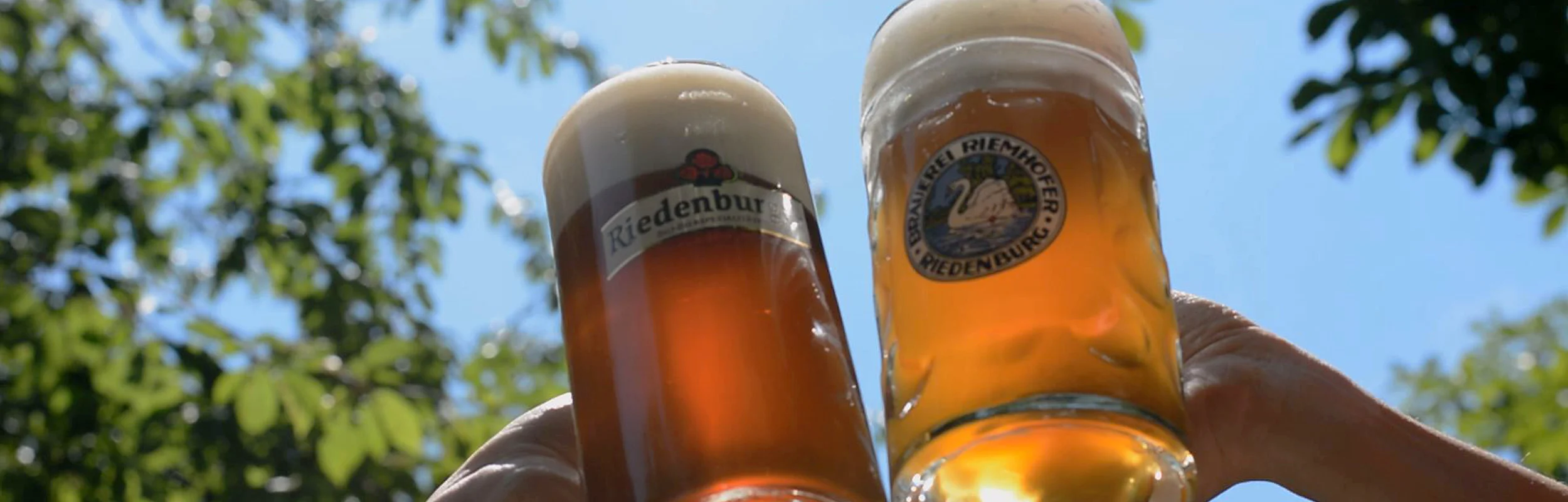 Zwei Personen mit Bierkrüge in der Hand stoßen im Sonnenschein an.