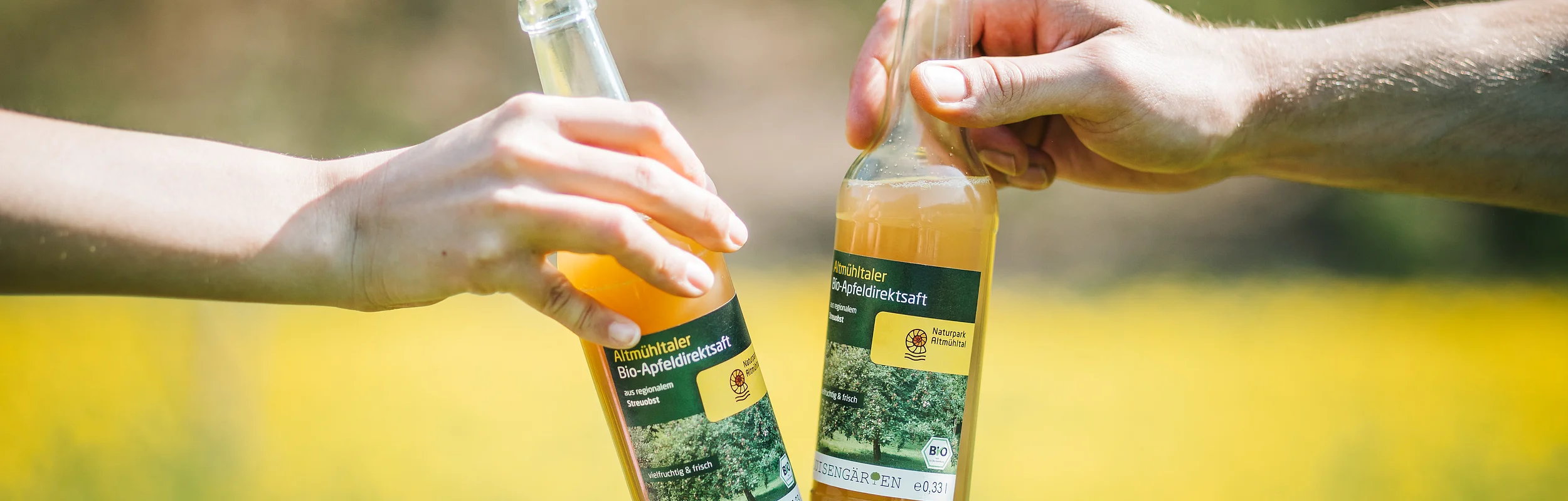 Zwei Personen stoßen mit zwei Altmühltaler Bio-Apfelsaft im Hintergrund die Sonnenstrahlen an.
